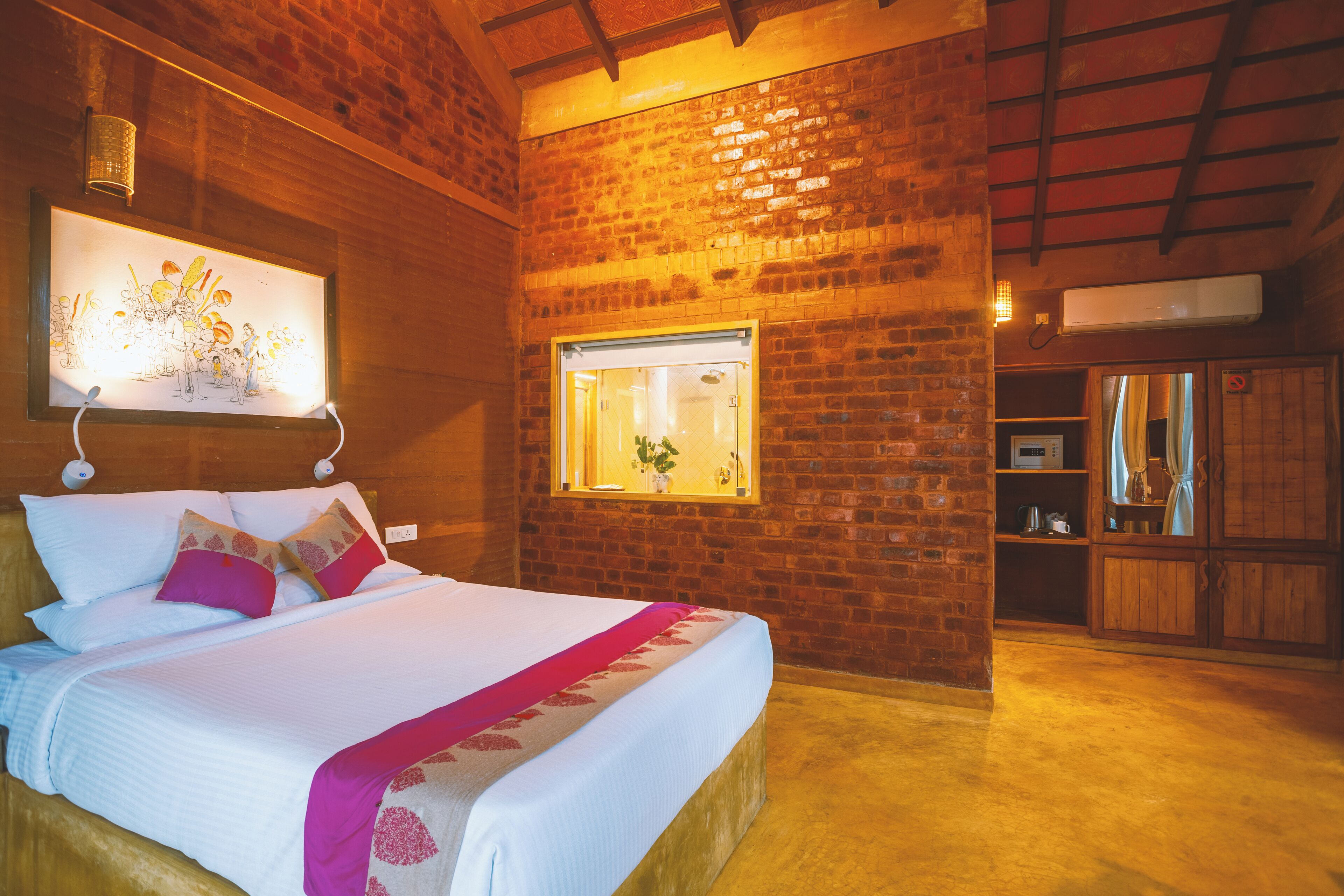 A hotel room with a brick wall, a bed with white bedding and pink accents, a framed artwork above the bed, a window with a view of greenery, and a kitchen area with wooden cabinets.