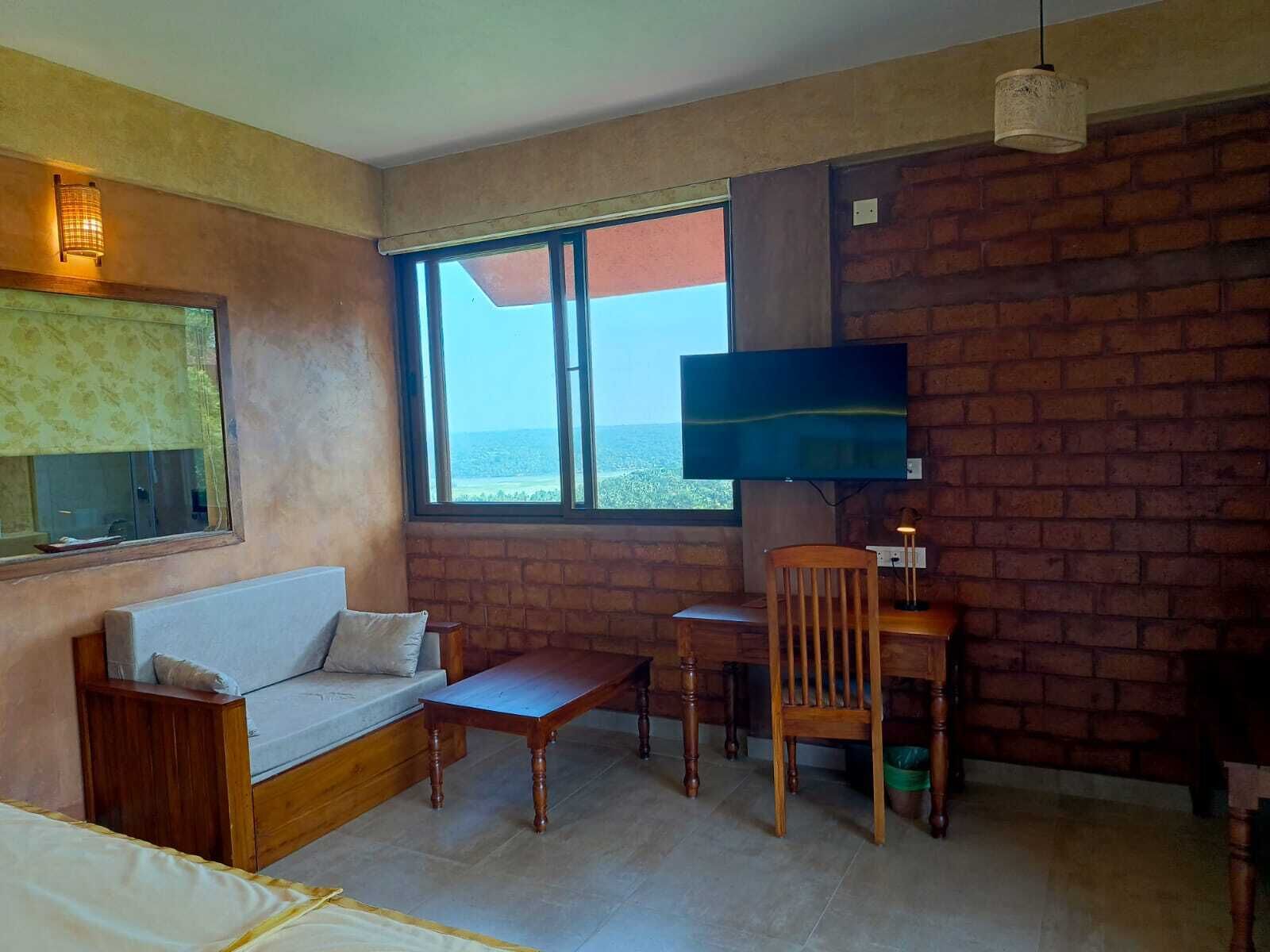 A room with a brick wall, a large window, a sofa, a desk with a chair, and a TV.