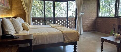 Habitación de hotel con una cama grande, una mesita de noche con teléfono, una pequeña mesa con planta y vistas a árboles a través de amplias ventanas.