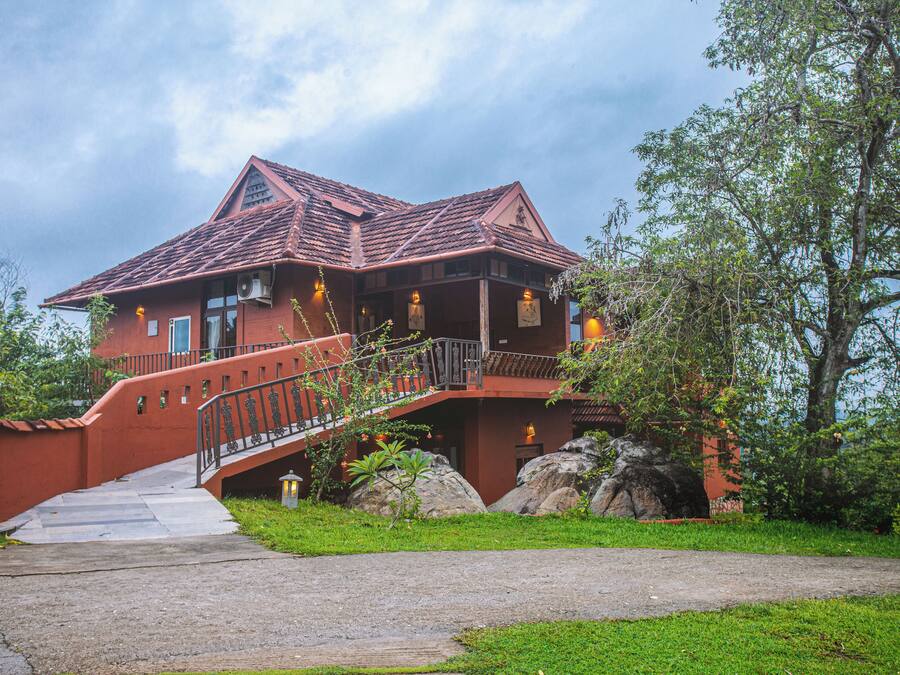 A large, red-roofed house with a staircase leading to a balcony, surrounded by greenery and rocks.