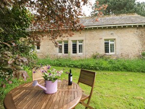 Una mesa de madera con un regador rosa y una botella de vino, una silla de madera, y vista a un jardín con un edificio de piedra y ventanas.