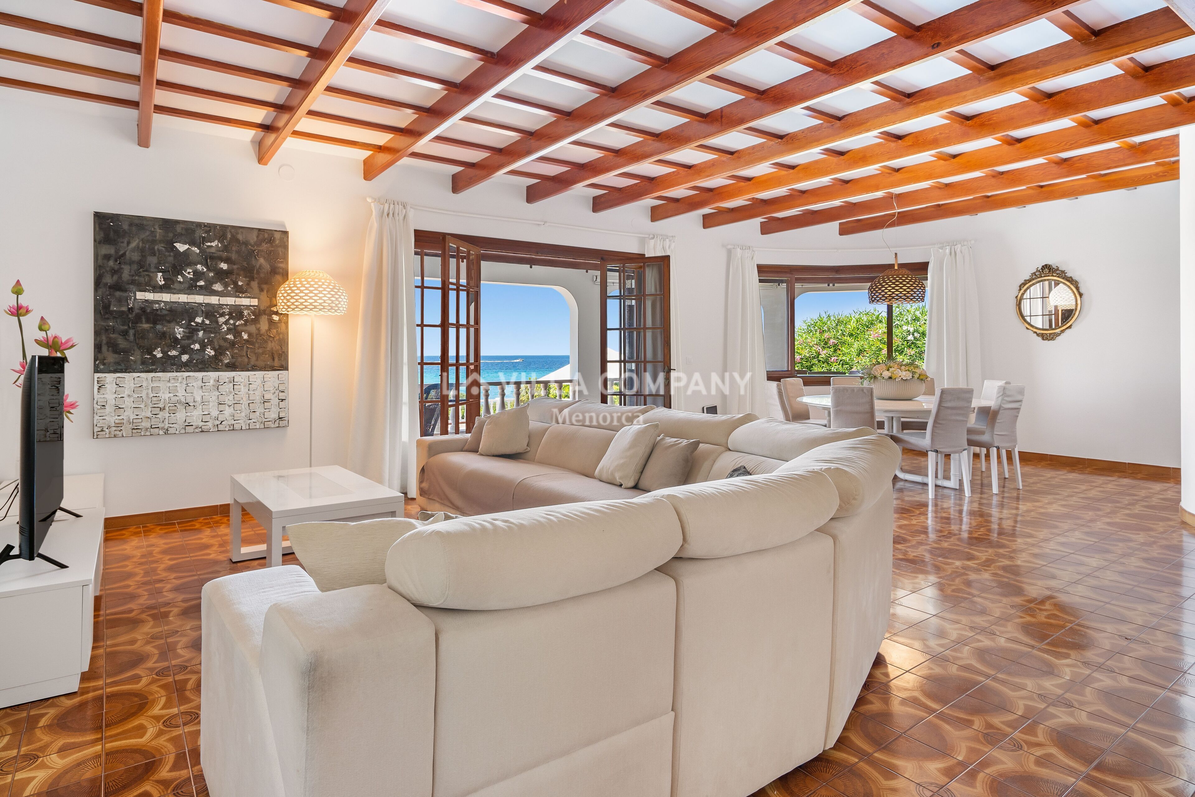 Un vaste espace de vie agrémenté d’un grand canapé beige, d’un parquet en bois et d’une vue sur la mer grâce à de grandes fenêtres ouvertes.
