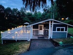 A small, single-story house with a porch and a deck, surrounded by trees.