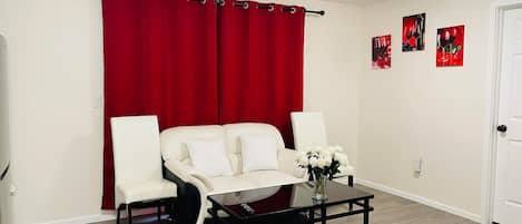 Une pièce avec un canapé blanc, deux fauteuils, une table basse en verre et un rideau rouge. On y trouve trois œuvres d’art murales rouges et une porte blanche.