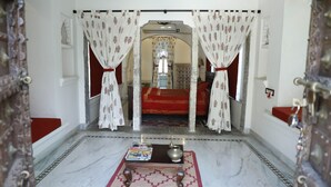 A hallway with a patterned rug, a small table with a book and a pot, and a red carpet leading to a room with a red bedspread. - Barabagh Deogarh (Devgarh)