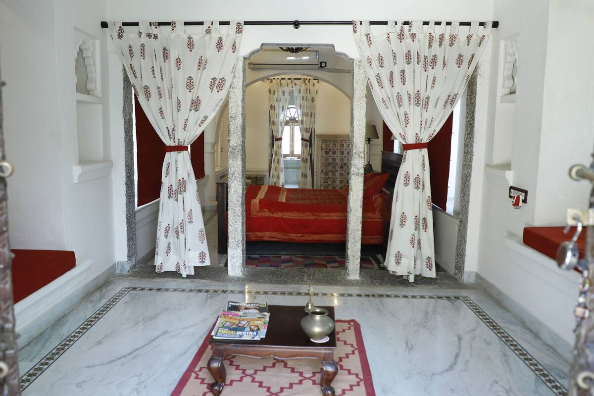 Um corredor com tapete estampado, uma mesinha com um livro e um vaso, e um tapete vermelho que leva a um quarto com colcha vermelha.