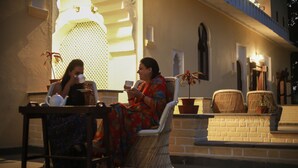 Two women seated at an outdoor table with tea cups, in front of a building with a balcony and potted plants. - Barabagh Deogarh (Devgarh)