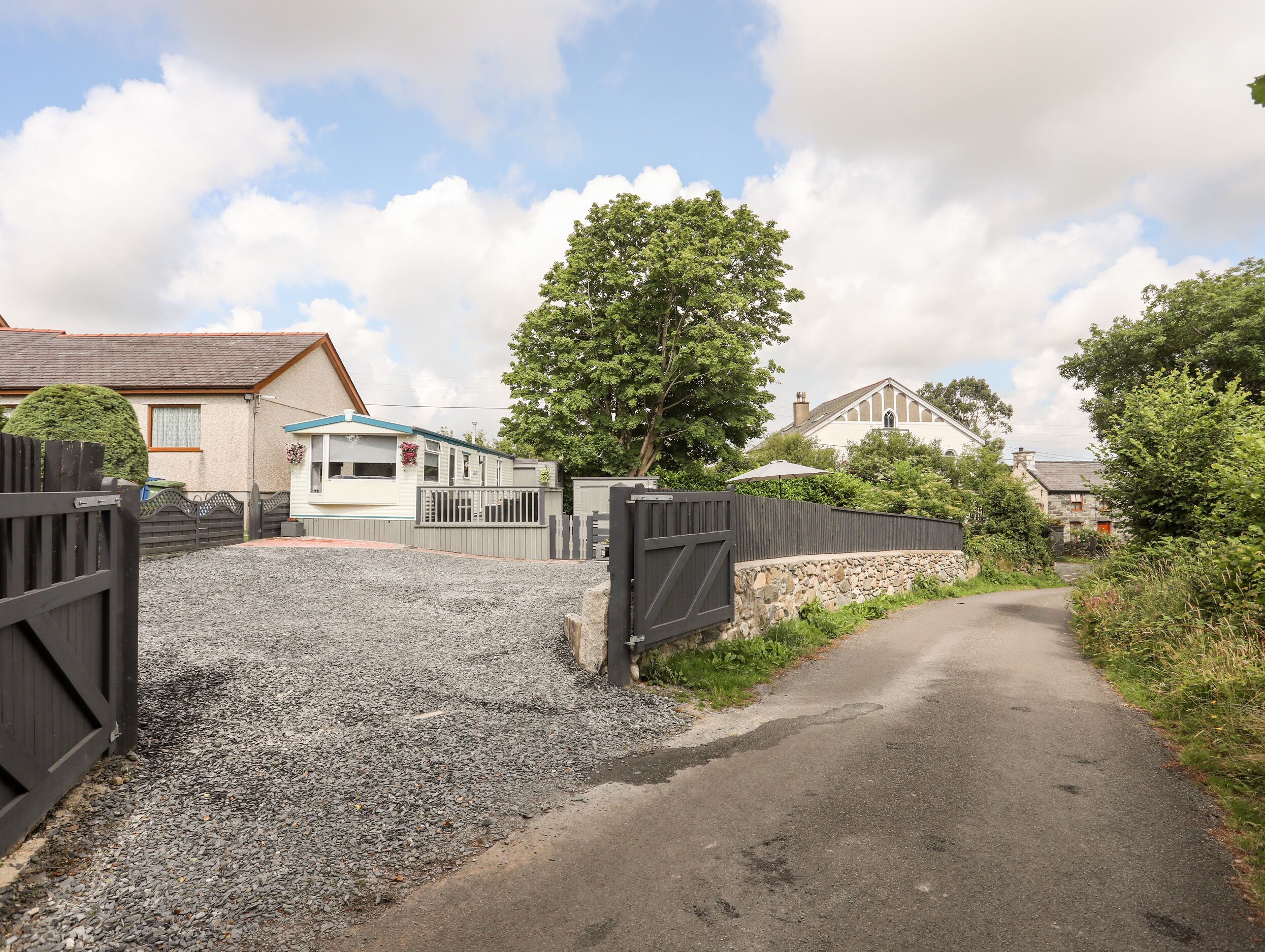 A gravel driveway leading to a gated entrance with a house and a large tree in the background.