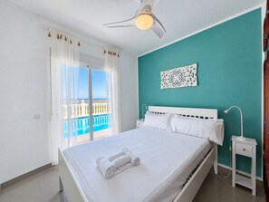 A bedroom with a bed, a nightstand, a ceiling fan, a view of a balcony with a pool, and a teal-colored wall. - Villa La Sella DJ with Private Pool and Golf Views (Pedreguer)