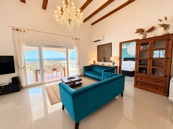A spacious living room with a large sofa, a television, and a chandelier. There is a balcony with a view of the sea. - Villa La Sella DJ with Private Pool and Golf Views (Pedreguer)