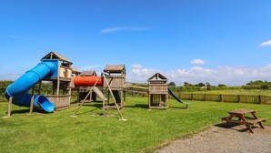 Un parque infantil con un tobogán azul, una estructura cilíndrica roja y casas de juego de madera.