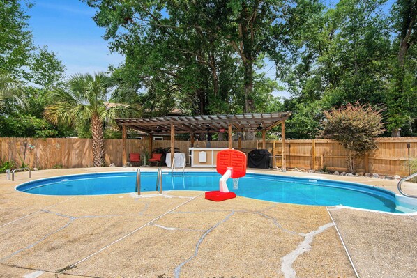 A swimming pool with a basketball hoop, surrounded by a wooden fence and trees.