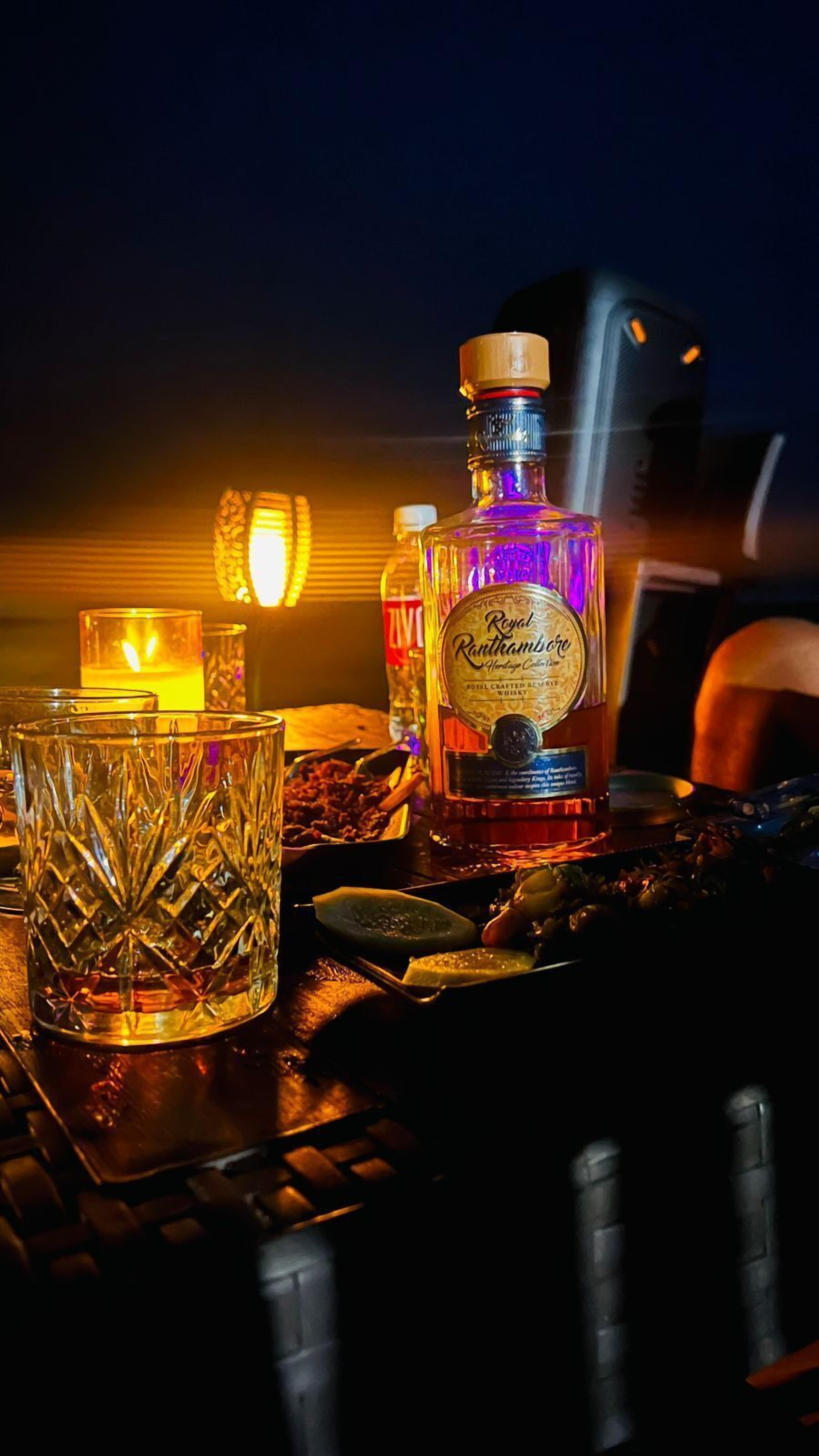 A bottle of Royal Ranthambore whisky, a glass with a textured design, and a lit candle on a table.