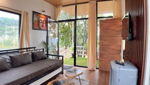 A modern living room with a sofa, a coffee table, and a TV. There is a sliding glass door leading to a balcony with a view of greenery. - Mango Forest Agro Tourism Resort (Pune)