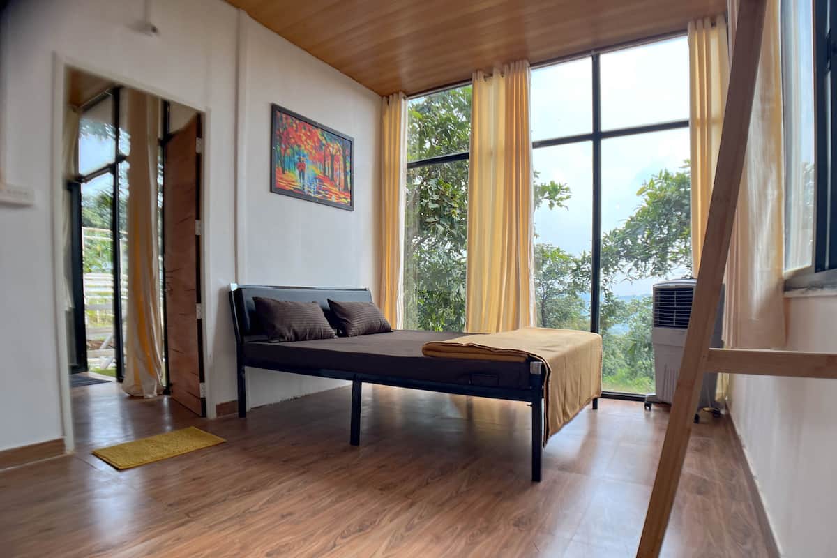 A bedroom with a bed, wooden floor, large windows, and a painting on the wall.