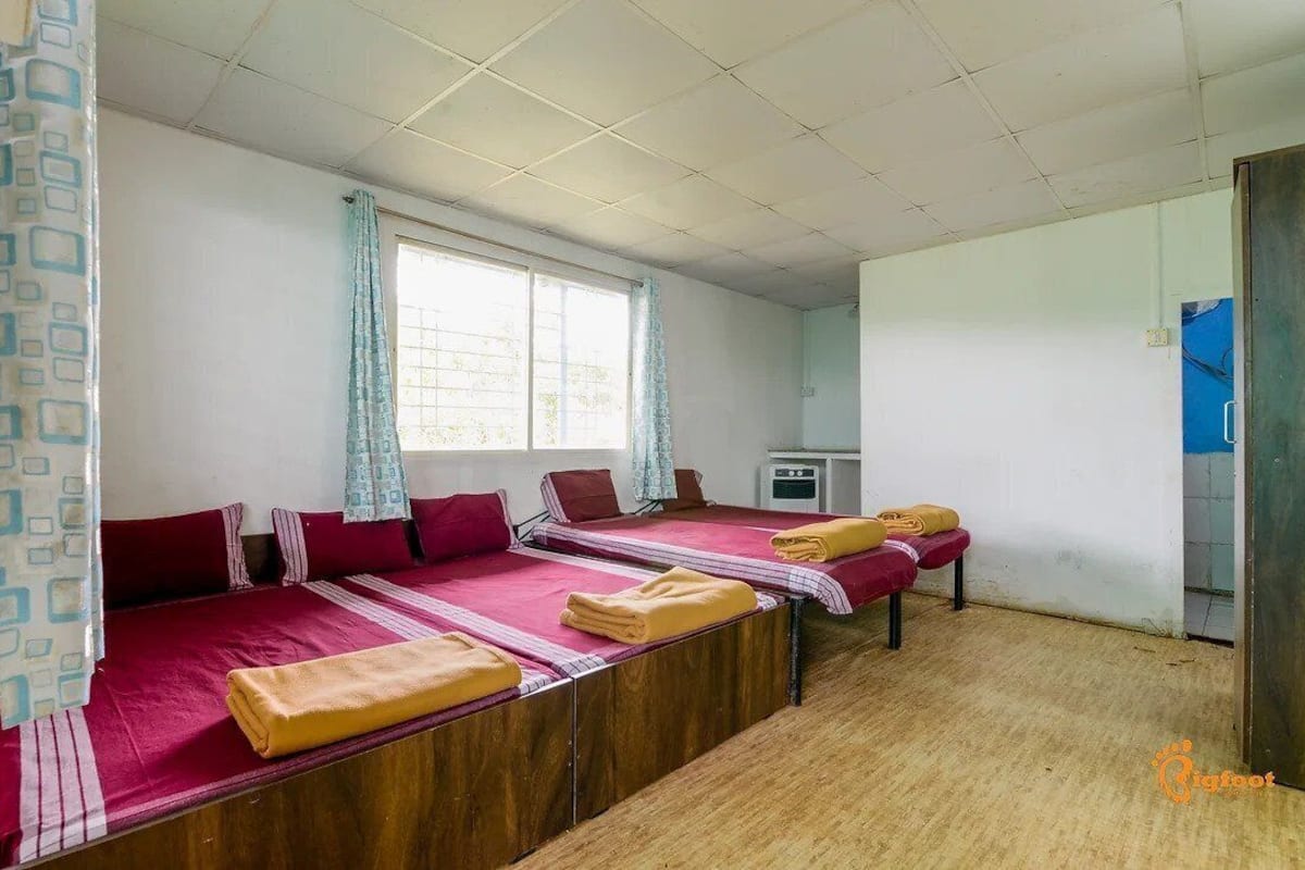 A room with three beds, a window with curtains, a small desk, and a wooden floor.