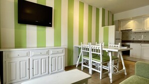 A room with a dining table and chairs, a television mounted on the wall, and a kitchen area with cabinets and a countertop.