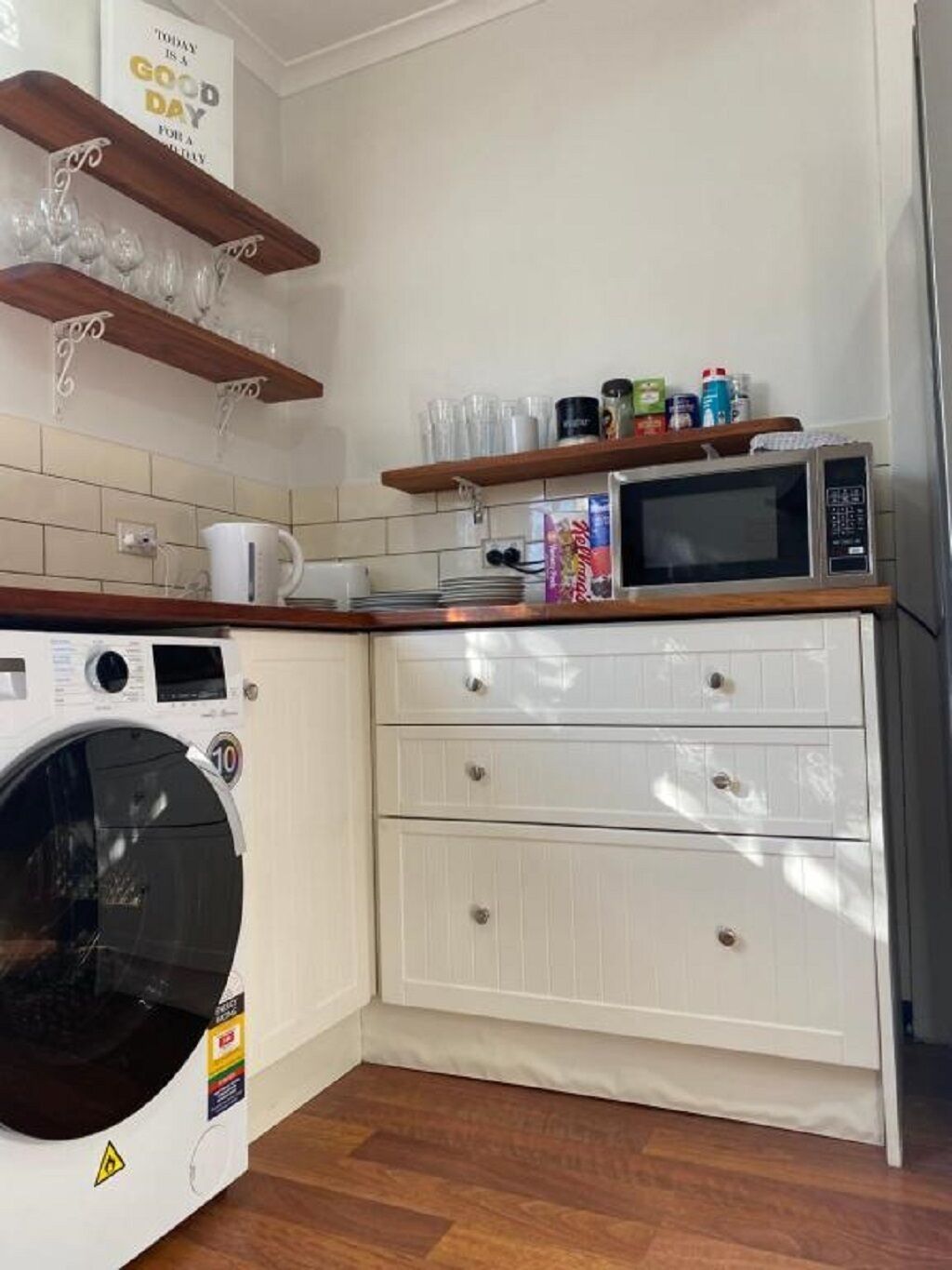 A compact kitchen with a washing machine, microwave, and wooden shelves.