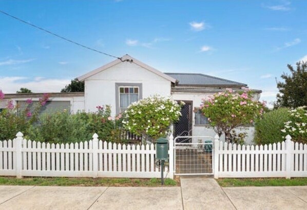 A single-story house with a white picket fence, a garage, and a small garden with pink flowers.