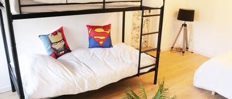 A bunk bed with two pillows, a small table with a plant, and a lamp in a room with wooden flooring and a white bed in the corner.
