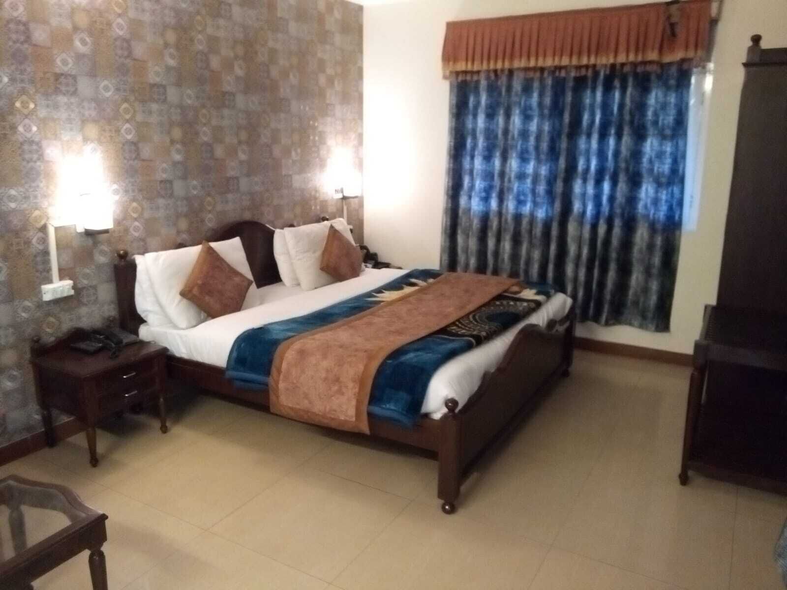 Una habitación de hotel con cama, mesitas de noche, lámpara, televisor y ventana con cortinas.