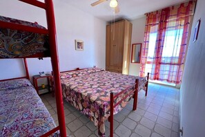 A room with bunk beds, a wooden wardrobe, a desk with a lamp, and a window with curtains.