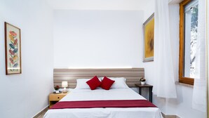 Ein ordentlich bezogenes Bett mit einem roten Akzentkissen, ein Nachttisch mit Lampe und ein gerahmtes Bild an der Wand.