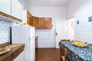 A compact kitchen with a refrigerator, wooden cabinets, a dining table with a floral tablecloth, and a wall with patterned tiles.