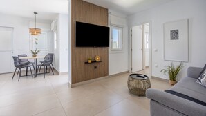A modern living room with a flat-screen TV, a dining area with a table and chairs, and a grey sofa. - Casita Meraki 3 with Heated Indoor Pool, Garden, Terrace, and Wi-Fi (Chiclana de la Frontera)