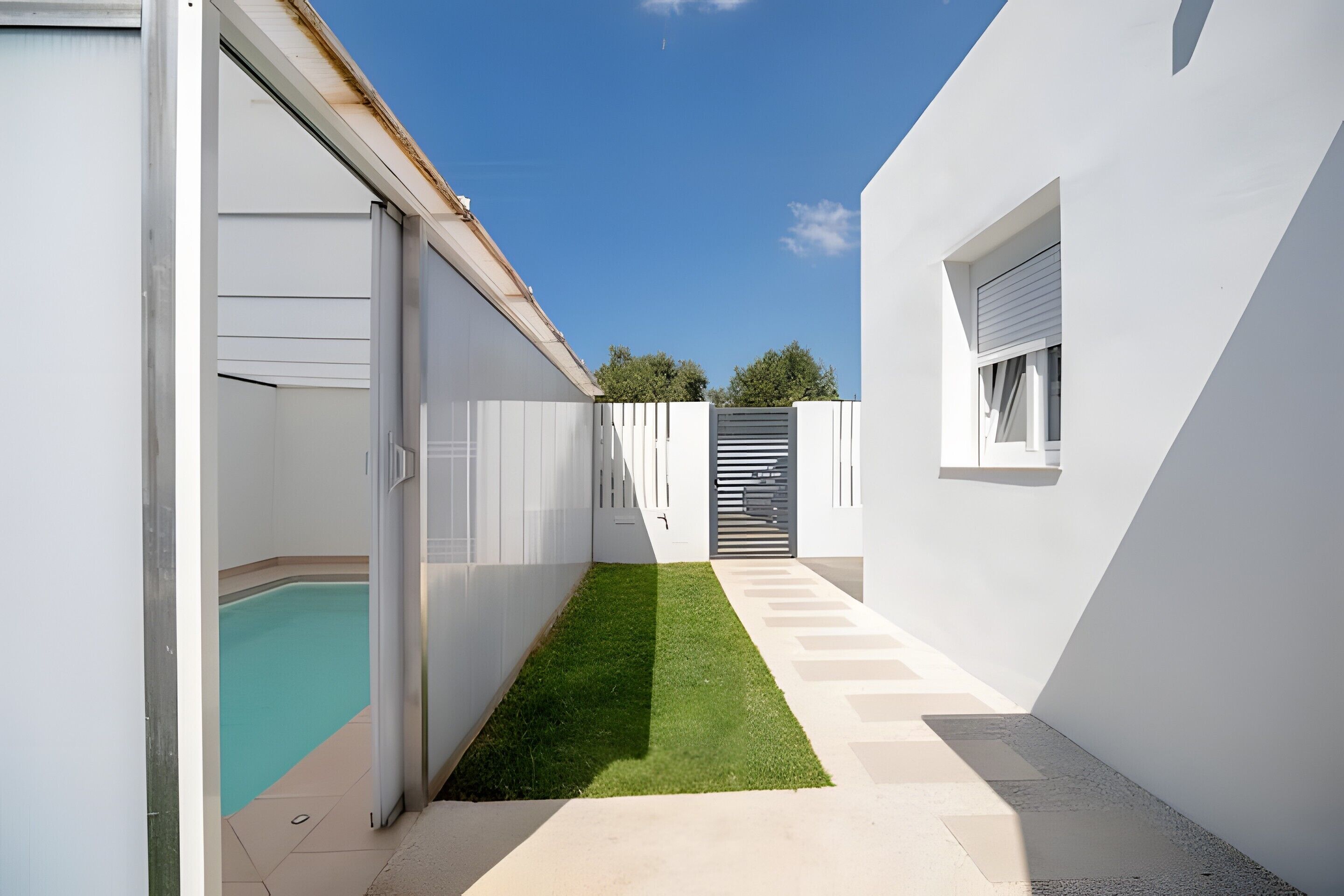 Un espacio exterior estrecho con piscina, césped verde y paredes blancas.