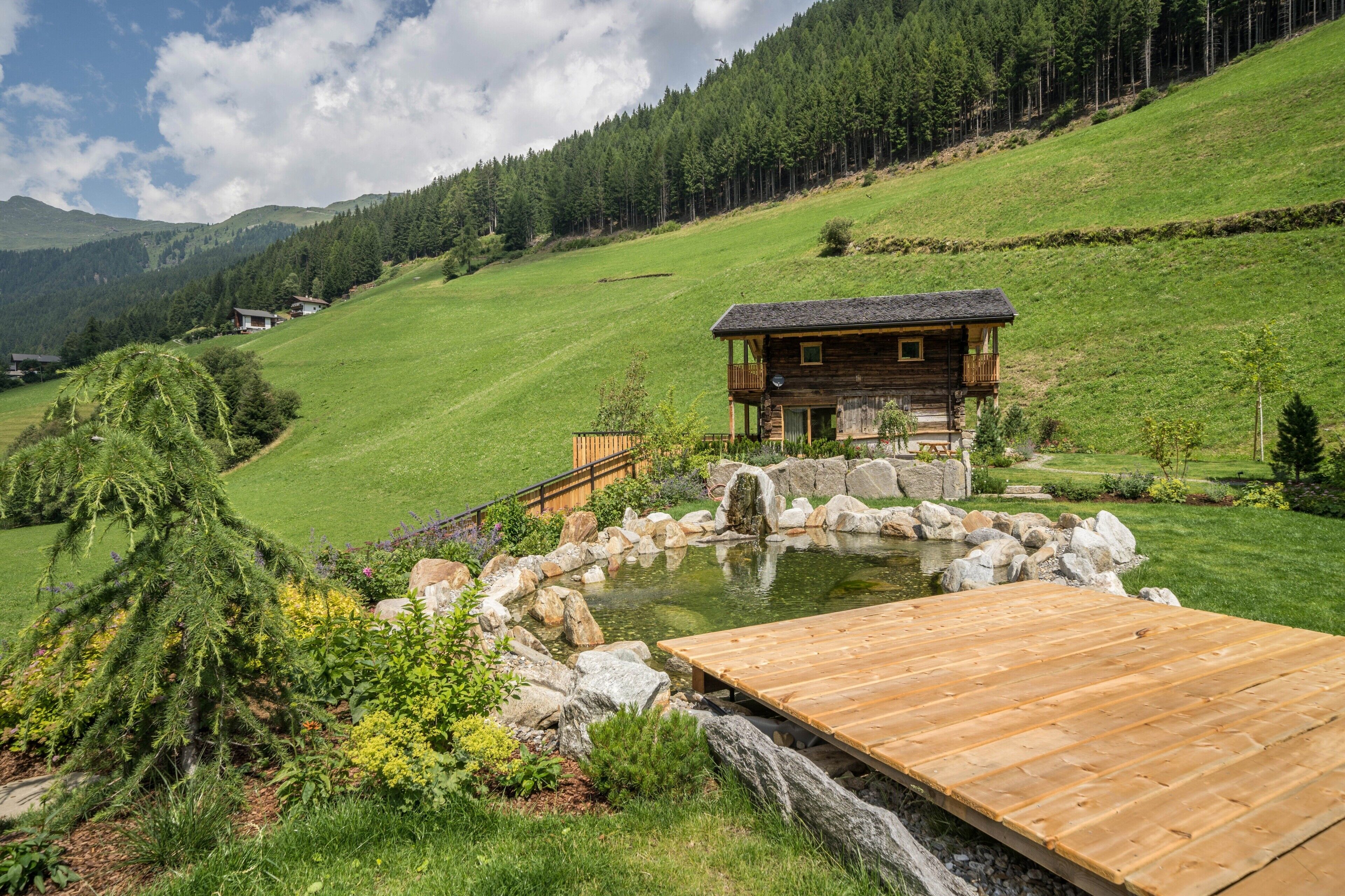Uma cabana de madeira com varanda, rodeada por um lago e vegetação, situada numa paisagem montanhosa.