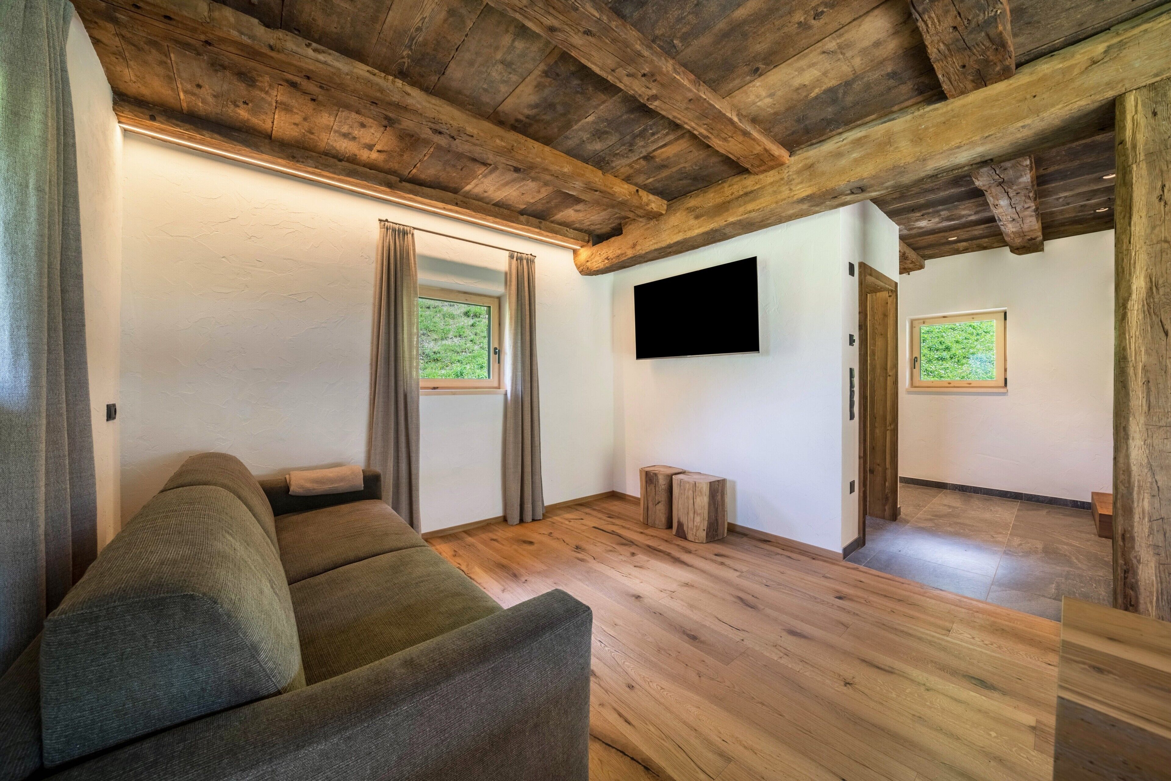 Una habitación con techo de madera, un sofá, un televisor de pantalla plana y dos ventanas con cortinas.