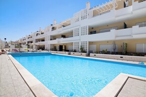 A swimming pool with a clear blue water, surrounded by a white building with balconies and a tiled deck.