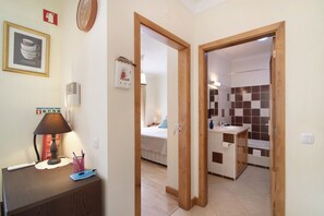 A hotel room with a wooden desk, a lamp, a framed picture, a bedside table, a bed, a bathroom with a sink and mirror, and a checkered tile backsplash.