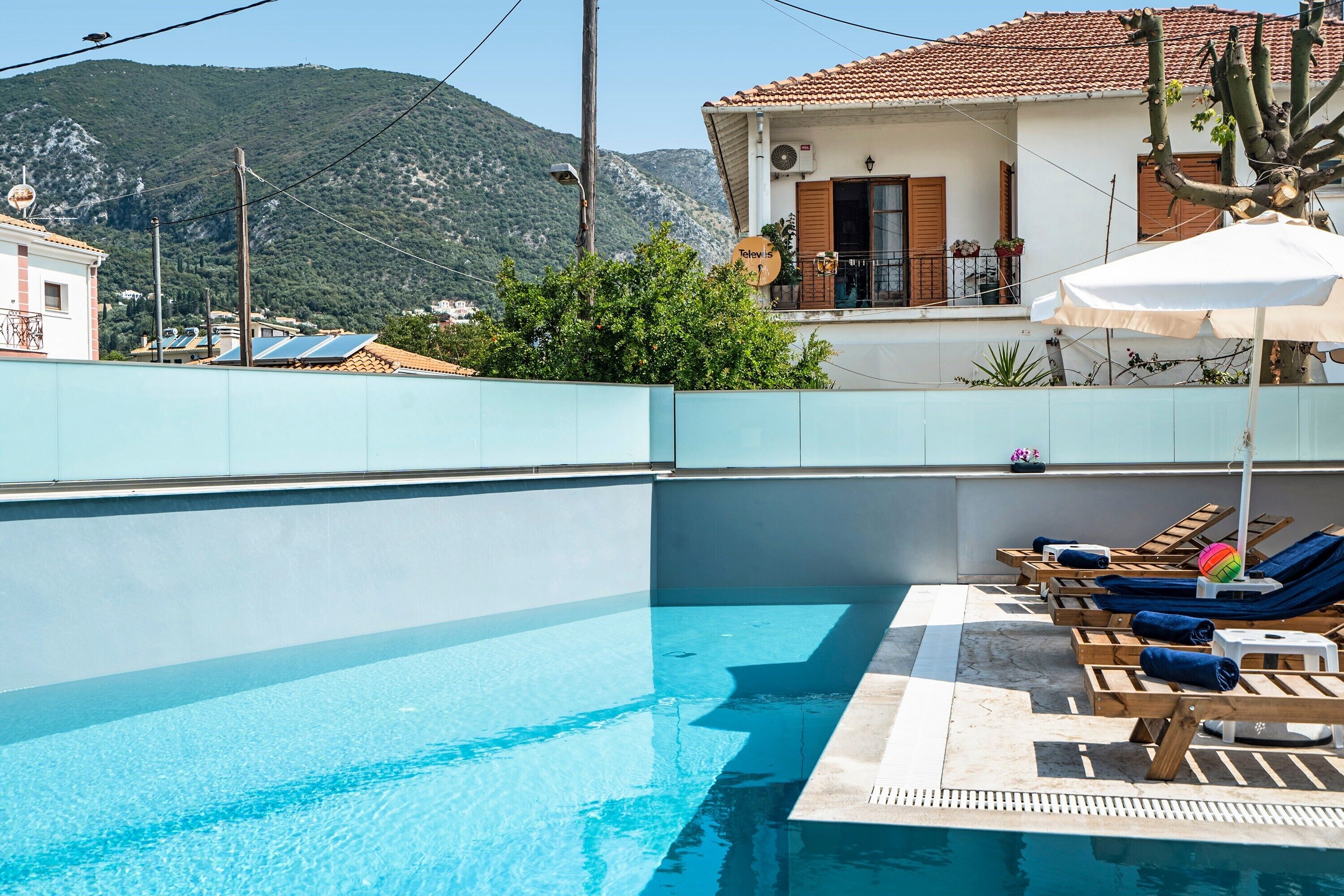 A swimming pool with lounge chairs and an umbrella, surrounded by a low wall and a building with a tiled roof.