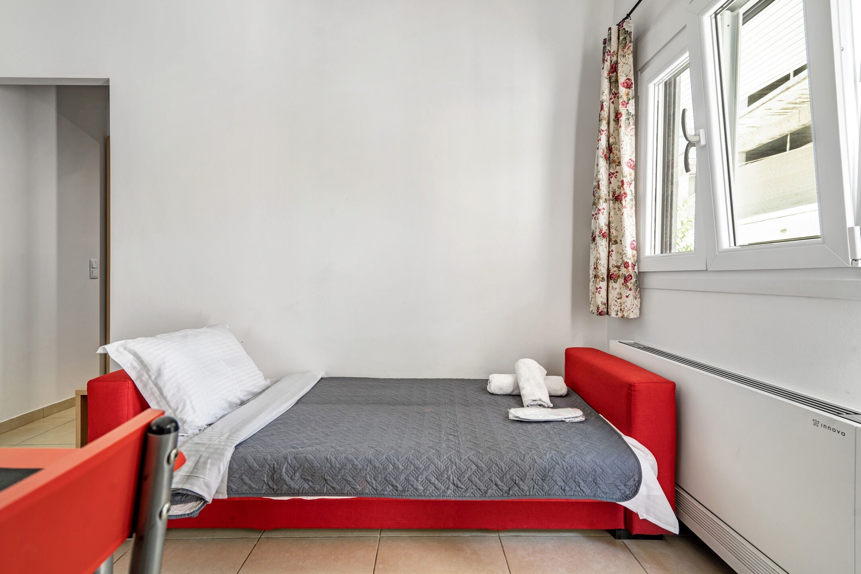 Een kleine eenpersoonskamer met een rode bedframe, een grijze spreidek en een wit kussen. Er is een raam met bloemen gordijnen, een radiator en een deur die naar een andere kamer leidt.