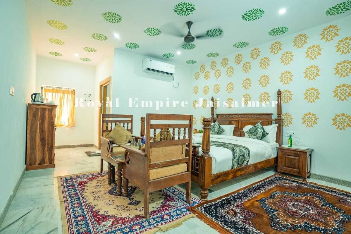 A bedroom with a four-poster bed, a wooden nightstand, a desk, and a chair. There is a ceiling fan, an air conditioning unit, and patterned carpets.