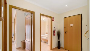 Un couloir d’hôtel avec des portes en bois, un lit visible au fond, une poubelle et un extincteur fixé au mur.