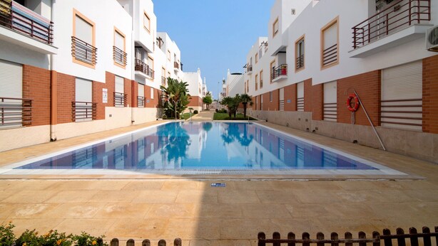 Une piscine entourée de bâtiments blancs dotés de balcons, un ciel dégagé et une bouée de sauvetage au bord de la piscine.
