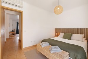 A bedroom with a bed, a bedside table, a hanging light fixture, and a view into another room.