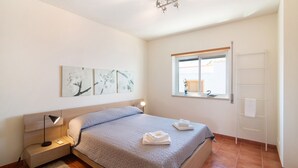 Un dormitorio con cama, mesita de noche, lámpara, obra de arte, ventana y un perchero para toallas.