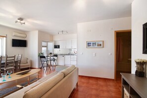 A modern living room with a sofa, dining table, and chairs, a kitchen with white cabinets, and a flat-screen TV mounted on the wall.