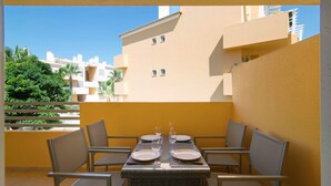 A dining area with a table set for four, surrounded by chairs, on a balcony with a view of neighboring buildings and greenery.
