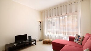 A living room with a flat-screen TV on a black stand, a red sofa, a small ottoman, and a floor lamp. - Apartment 'Royal Cabanas Golf 88' with Shared Pool, Shared Garden and Air Conditioning (Cabanas de Tavira)