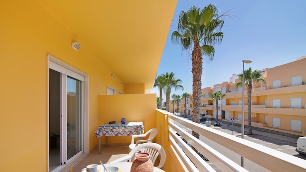 A balcony with a table, chairs, a vase, and a view of palm trees and buildings. - Apartment 'Royal Cabanas Golf 88' with Shared Pool, Shared Garden and Air Conditioning (Cabanas de Tavira)