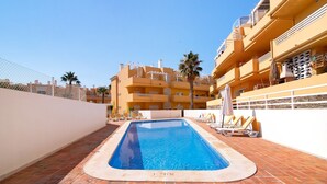 A swimming pool with a brick deck, surrounded by lounge chairs and umbrellas, with a building in the background. - Apartment 'Royal Cabanas Golf 88' with Shared Pool, Shared Garden and Air Conditioning (Cabanas de Tavira)