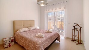 Une chambre à coucher comprenant un lit, un luminaire suspendu, une fenêtre avec des rideaux et un porte-manteau en bois.