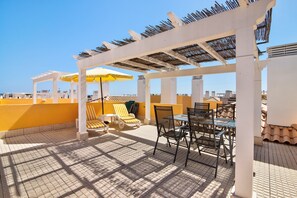 A rooftop terrace with sun loungers, a table with chairs, and a pergola structure.