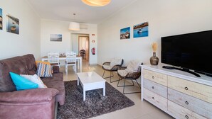 Een woonkamer met een bank, salontafel, televisie en rieten stoelen. In de achtergrond is een eethoek te zien.