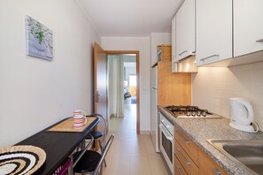 Cocina compacta con estufa a gas, microondas y cafetera. Se ve una mesa de comedor con una taza a rayas y un mantel tejido.